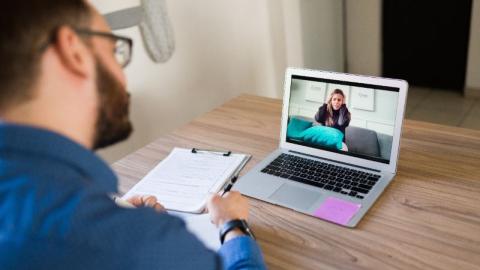 A woman sitting on a couch is pictured on a laptop screen talking to a man seated at a desk in an office.