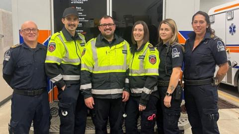 Six uniformed paramedics stand side by side in front of the back doors on an ambulance.