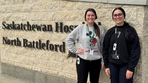 Two women who work as a registered psychiatric nurse and a mental health correctional officer stand in front of a sign on a brick wall that reads “Saskatchewan Hospital North Battleford”.