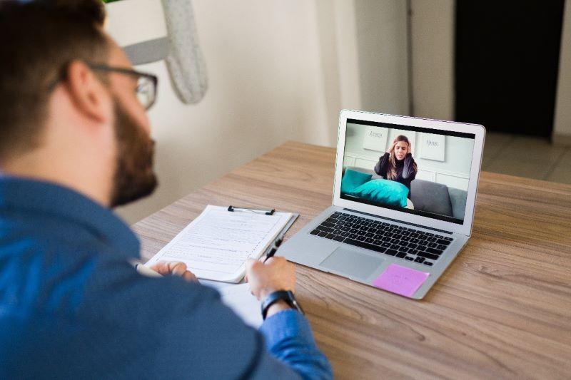 A woman sitting on a couch is pictured on a laptop screen talking to a man seated at a desk in an office.