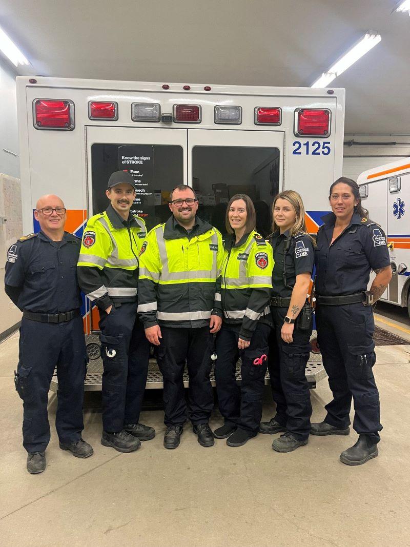 Six uniformed paramedics stand side by side in front of the back doors on an ambulance.